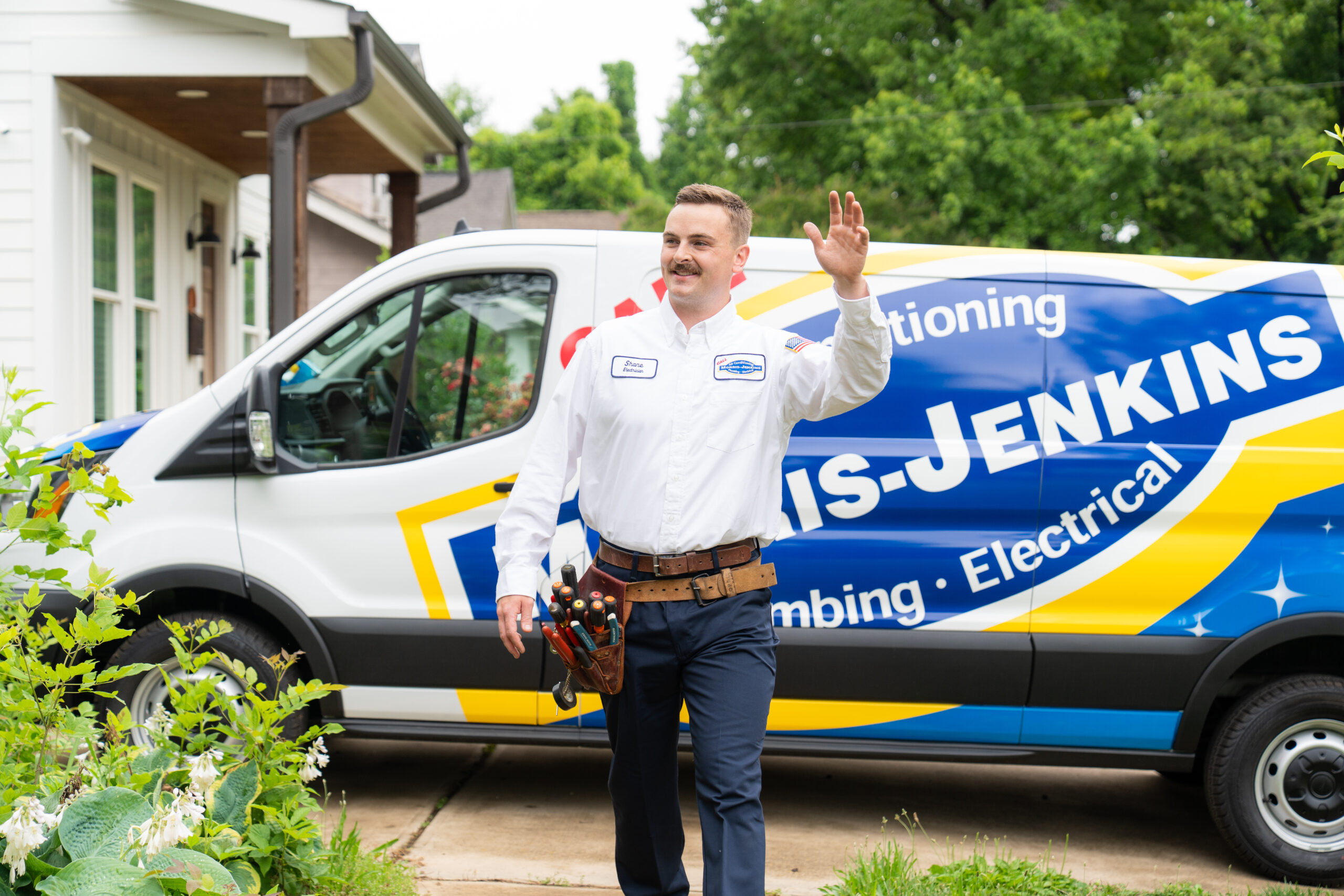 A technician wearing a tool belt waves while walking in front of a Jenkins van labeled for HVAC, plumbing, and electrical services in a residential area.