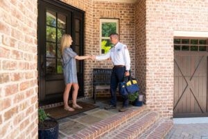 A woman stands at her front door shaking hands with a uniformed technician holding a tool bag on the porch of a brick house.