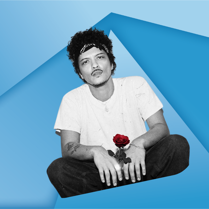 A person with curly hair, wearing a white t-shirt and dark pants, sits cross-legged holding a red rose, against a blue geometric background.