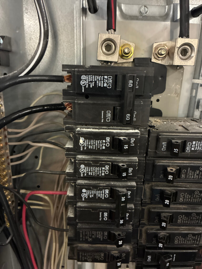 Close-up view of an electrical breaker panel with several circuit breakers, wires, and bus bars visible. Two heavy-gauge black wires are connected at the top.