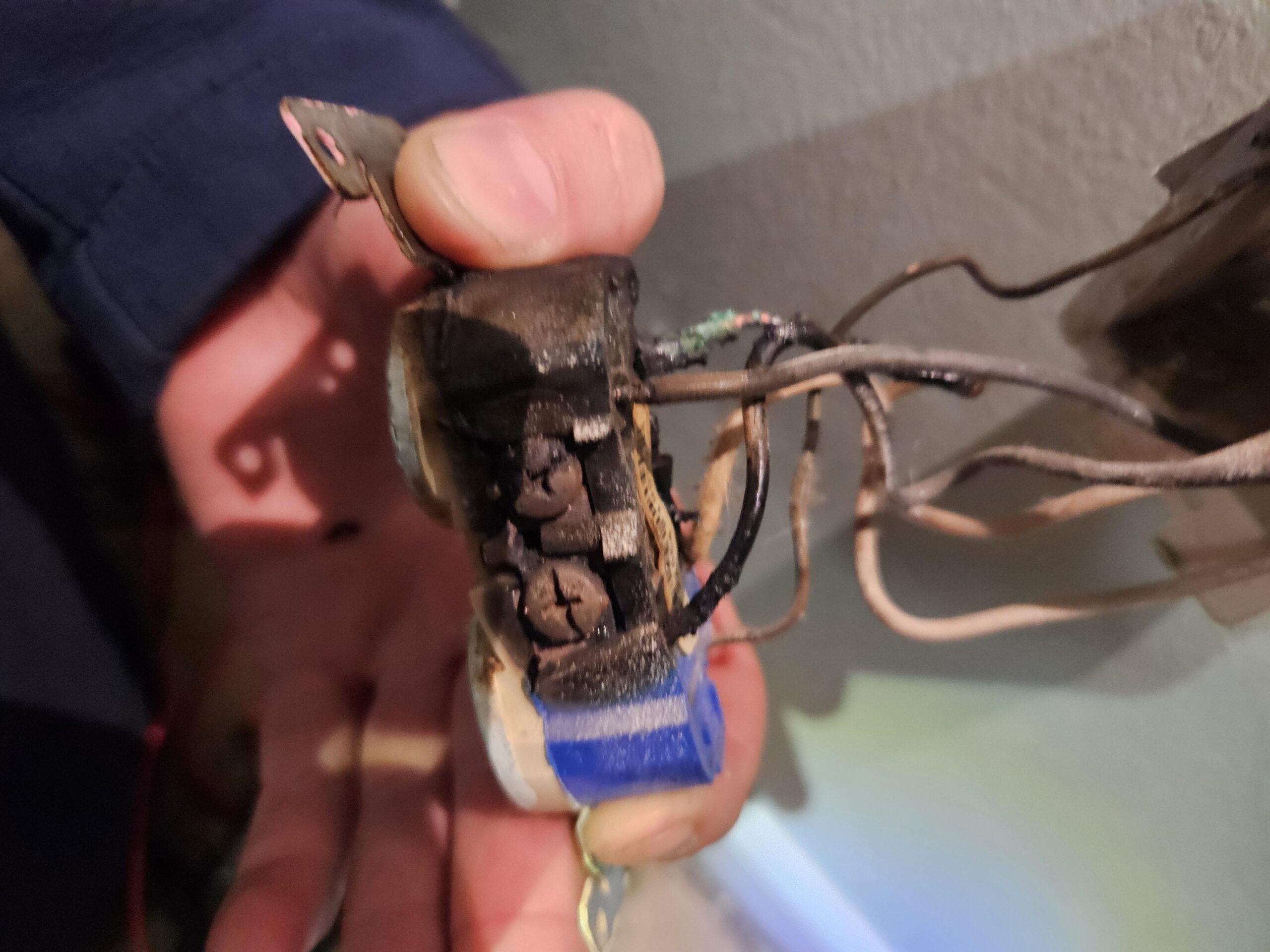A person's hand holds a burnt, damaged electrical outlet with exposed wires near a wall, indicating electrical failure or fire hazard.