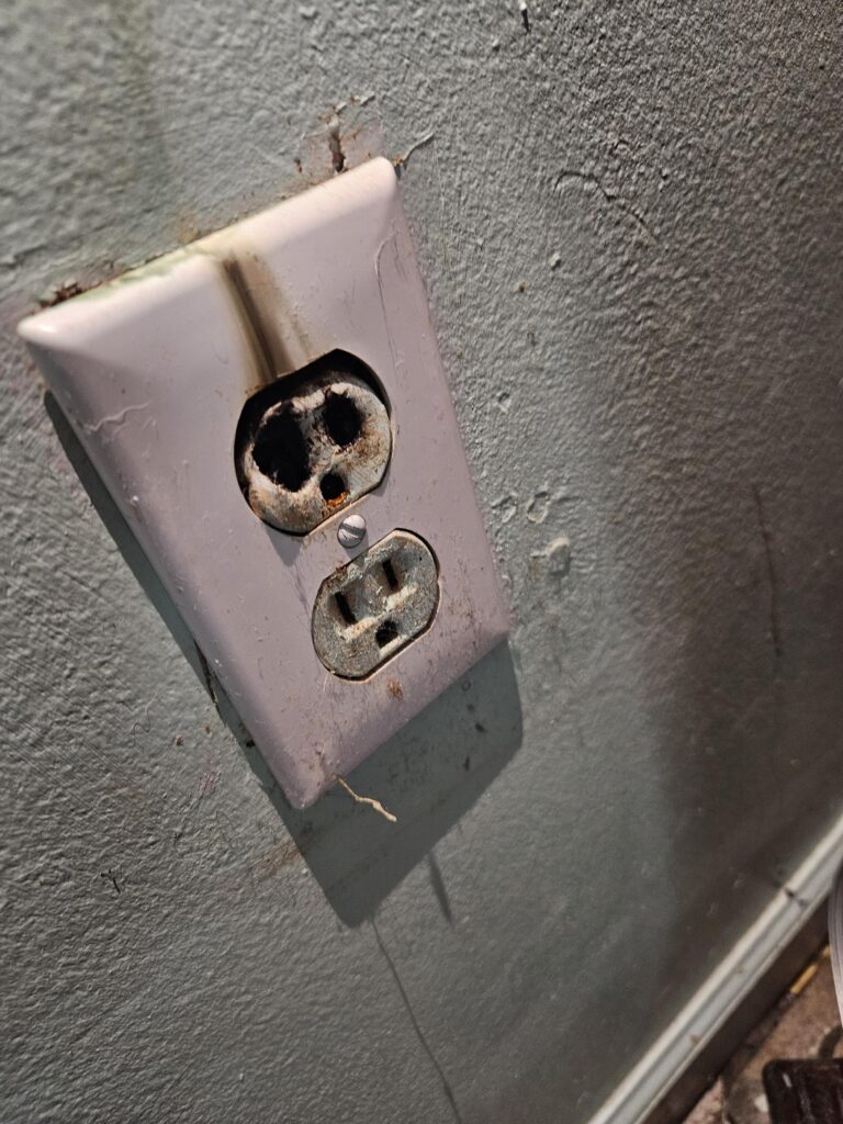 A dirty, damaged electrical outlet with burn marks is mounted on a stained wall near the baseboard.