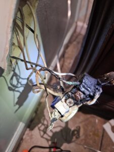 Close-up of a damaged electrical outlet with burnt and exposed wires protruding from a wall, showing signs of overheating and electrical failure.