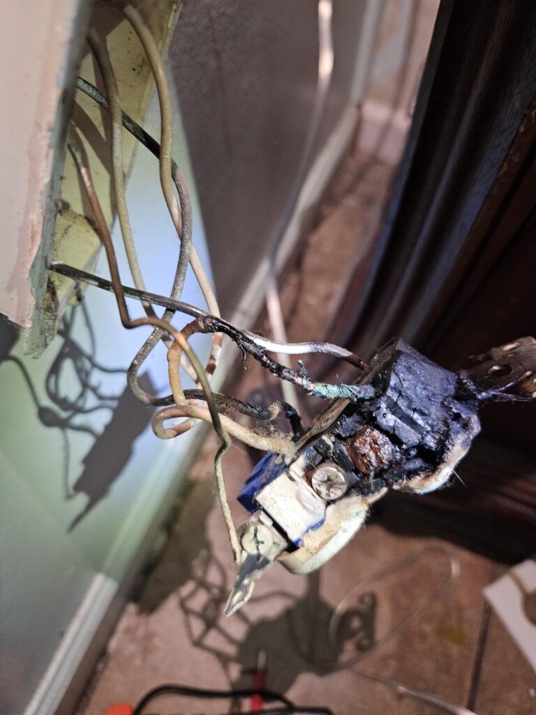 Close-up of a damaged electrical outlet with burnt and exposed wires protruding from a wall, showing signs of overheating and electrical failure.