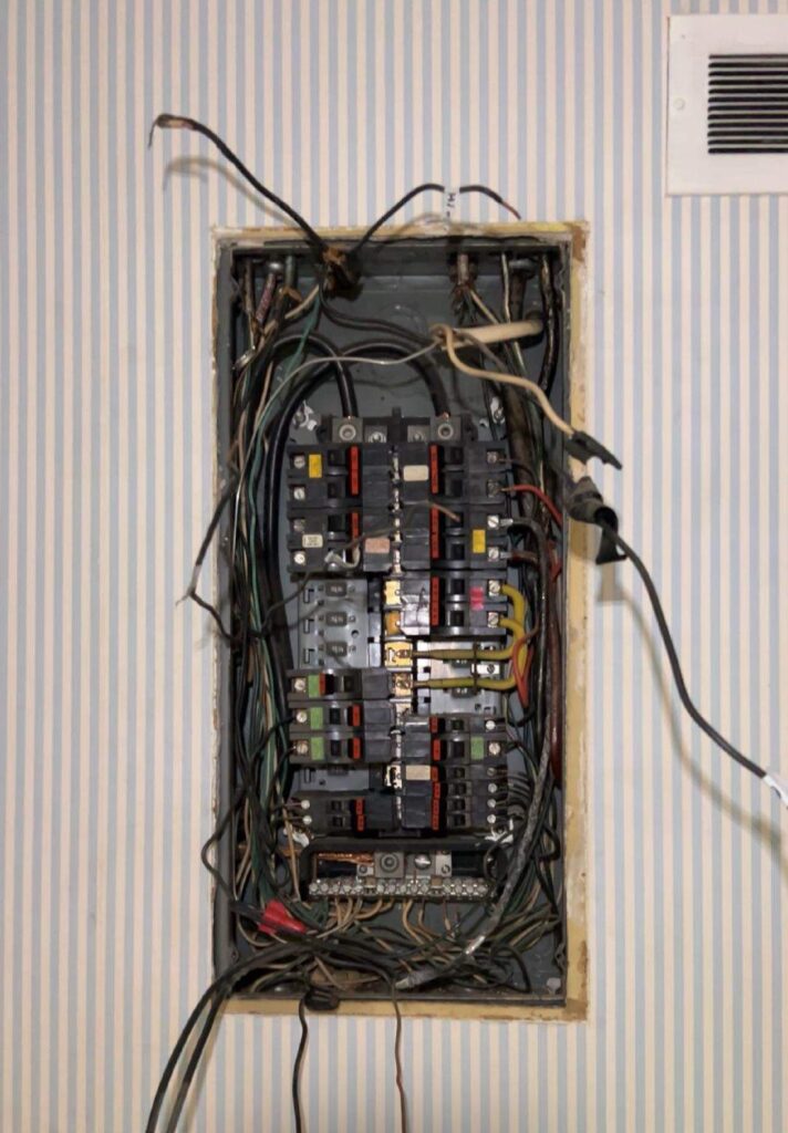 An open electrical breaker panel with exposed and tangled wires set in a wall with vertical striped wallpaper.