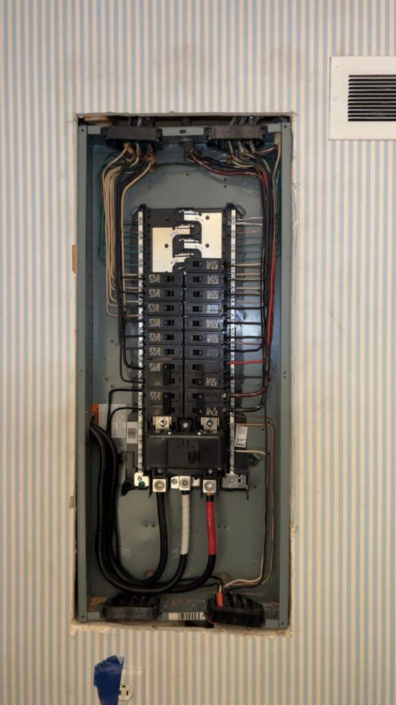 An open electrical panel with labeled circuit breakers, various colored wires, and a striped wallpaper background.