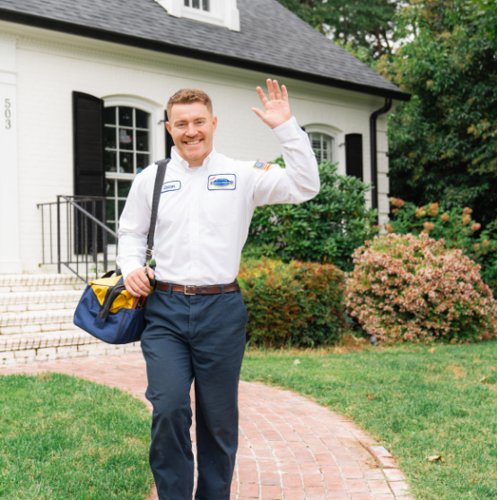 A smiling technician in a uniform waves while walking on a brick path toward a white house, carrying a blue bag.