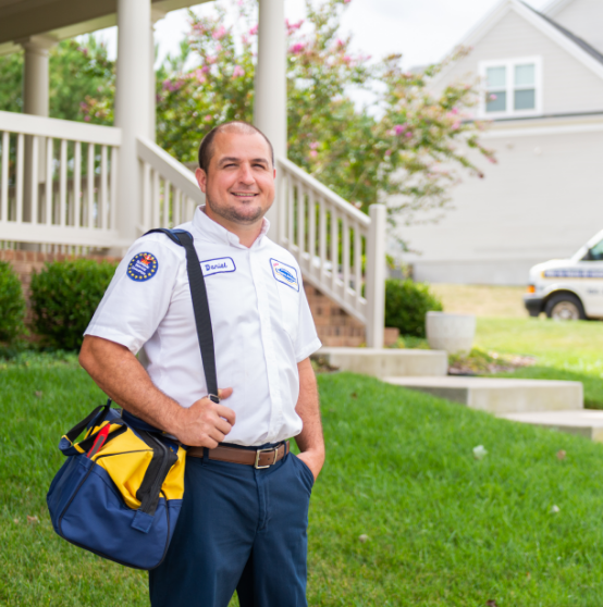 A male technician in a white uniform stands outside a house, holding a tool bag, with a service van parked in the background.