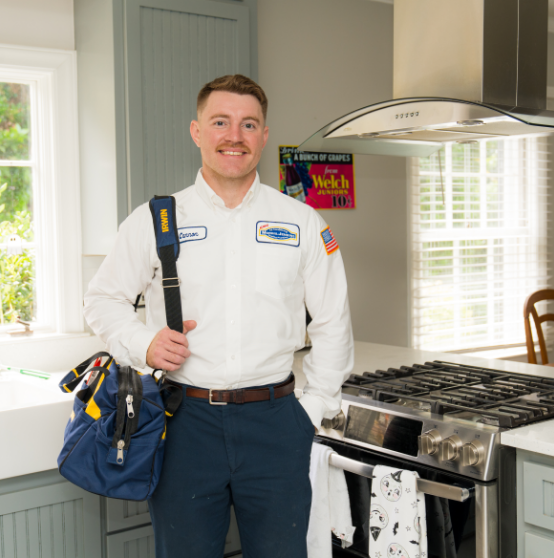 A man in a work uniform stands in a kitchen beside a stove, holding a tool bag and smiling at the camera.