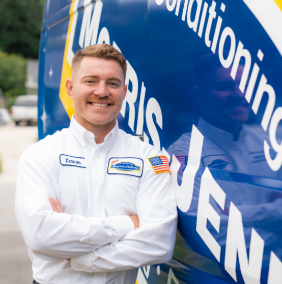 A man in a white work shirt with a name tag stands smiling with arms crossed in front of a blue service van with large lettering.