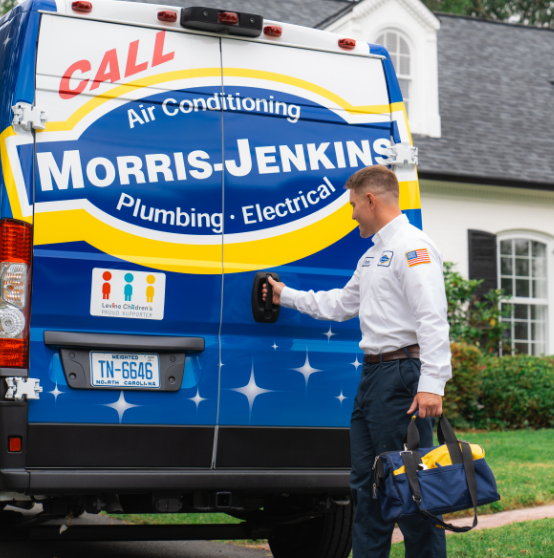 A technician stands by a Morris-Jenkins service van, holding a tool bag and reaching for the door handle outside a residential house.