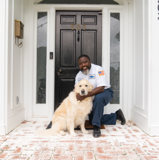 A man in a uniform kneels and hugs a large golden retriever on the front step of a house with a black door numbered 503.