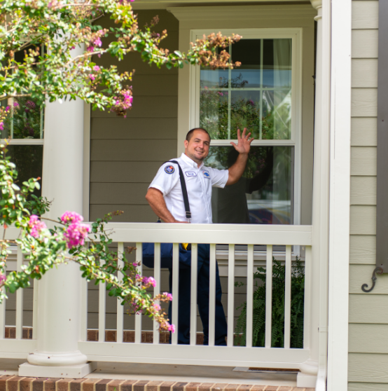 A man in a uniform stands on a front porch, smiling and waving, with flowering shrubs in the foreground.
