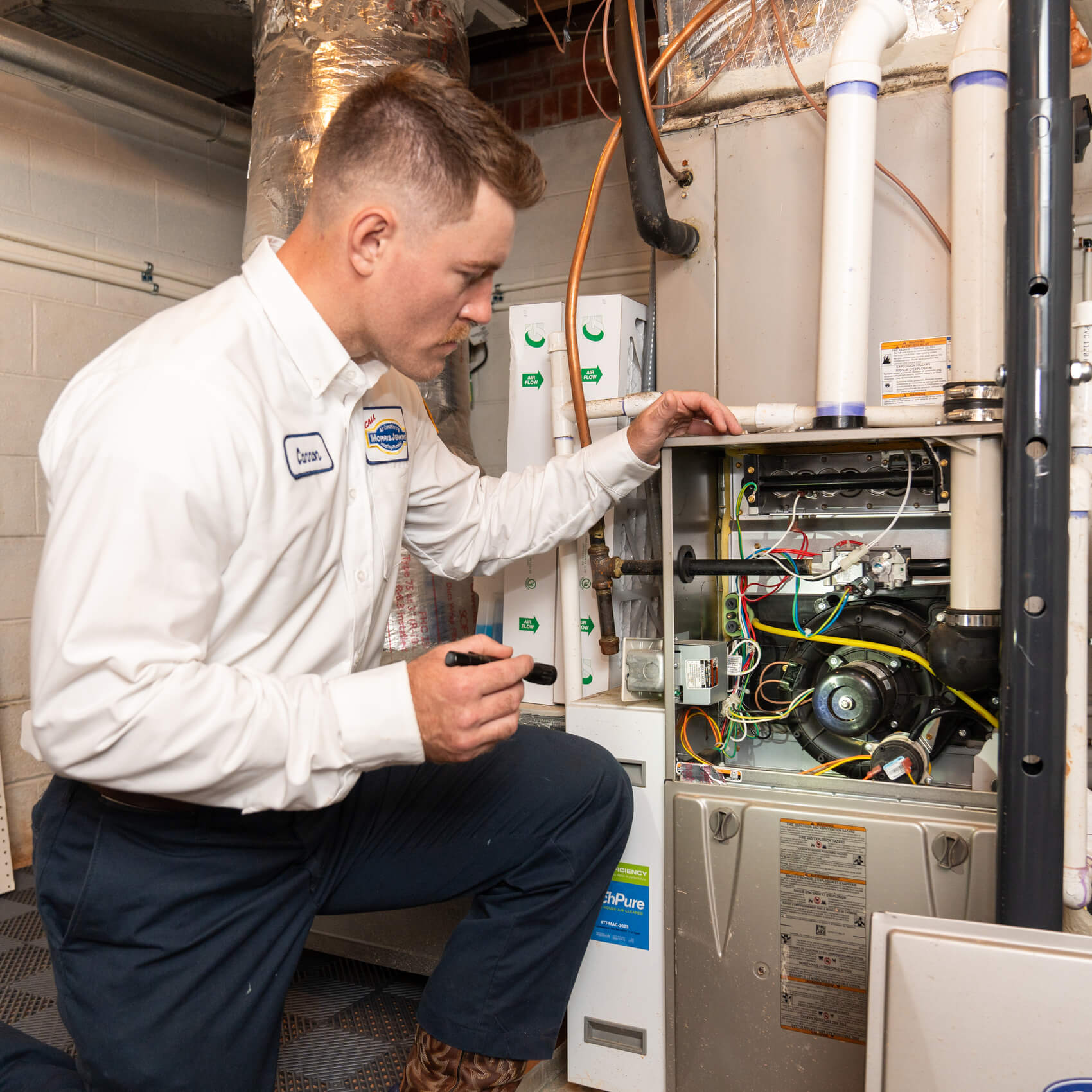 A technician inspects the interior components of a furnace in a utility room, holding a flashlight and wearing a uniform.