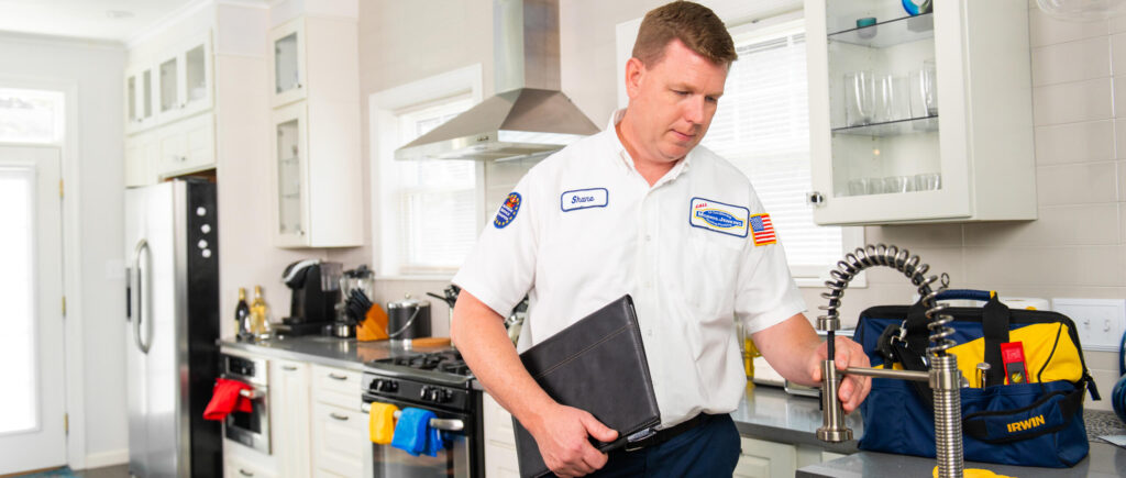 A plumber in uniform inspects a kitchen faucet while holding a tool case, with plumbing tools and bags on the counter nearby.