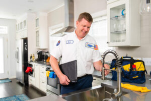 A plumber in uniform checks a kitchen faucet while holding a folder. A tool bag and cleaning cloth are on the countertop in a modern, well-lit kitchen.