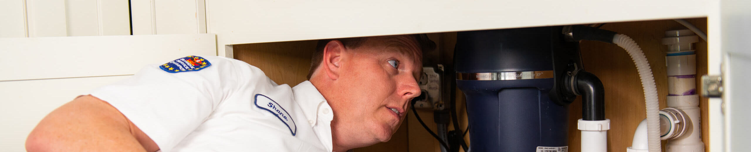A plumber in uniform examines plumbing and a garbage disposal unit under a kitchen sink.