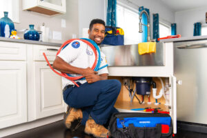 A plumber kneels in front of an open kitchen sink cabinet, smiling while holding red and blue hoses, with tools and bags nearby.