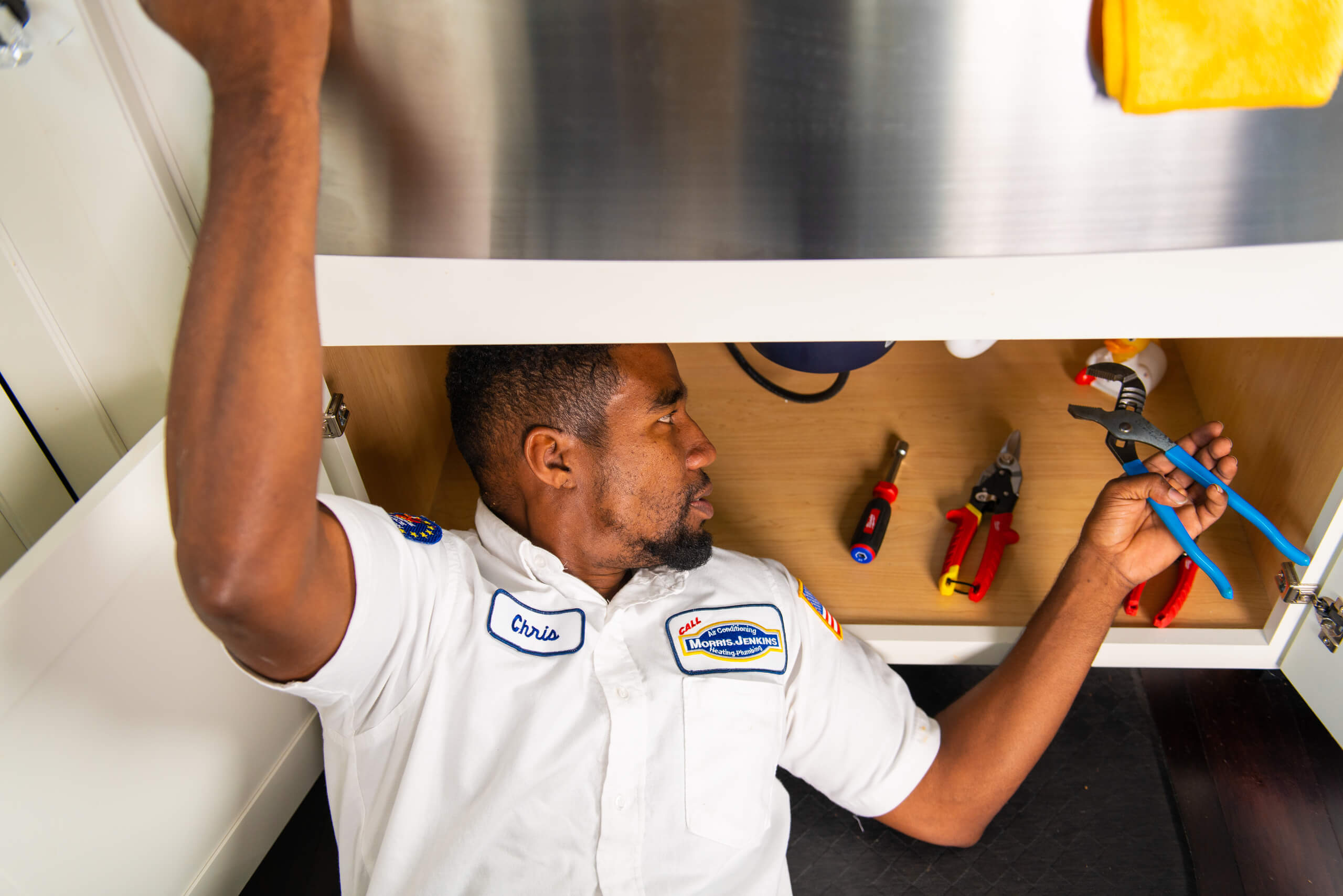 A plumber wearing a uniform lies under a sink, using pliers to work on plumbing, with various tools visible beside him inside the cabinet.