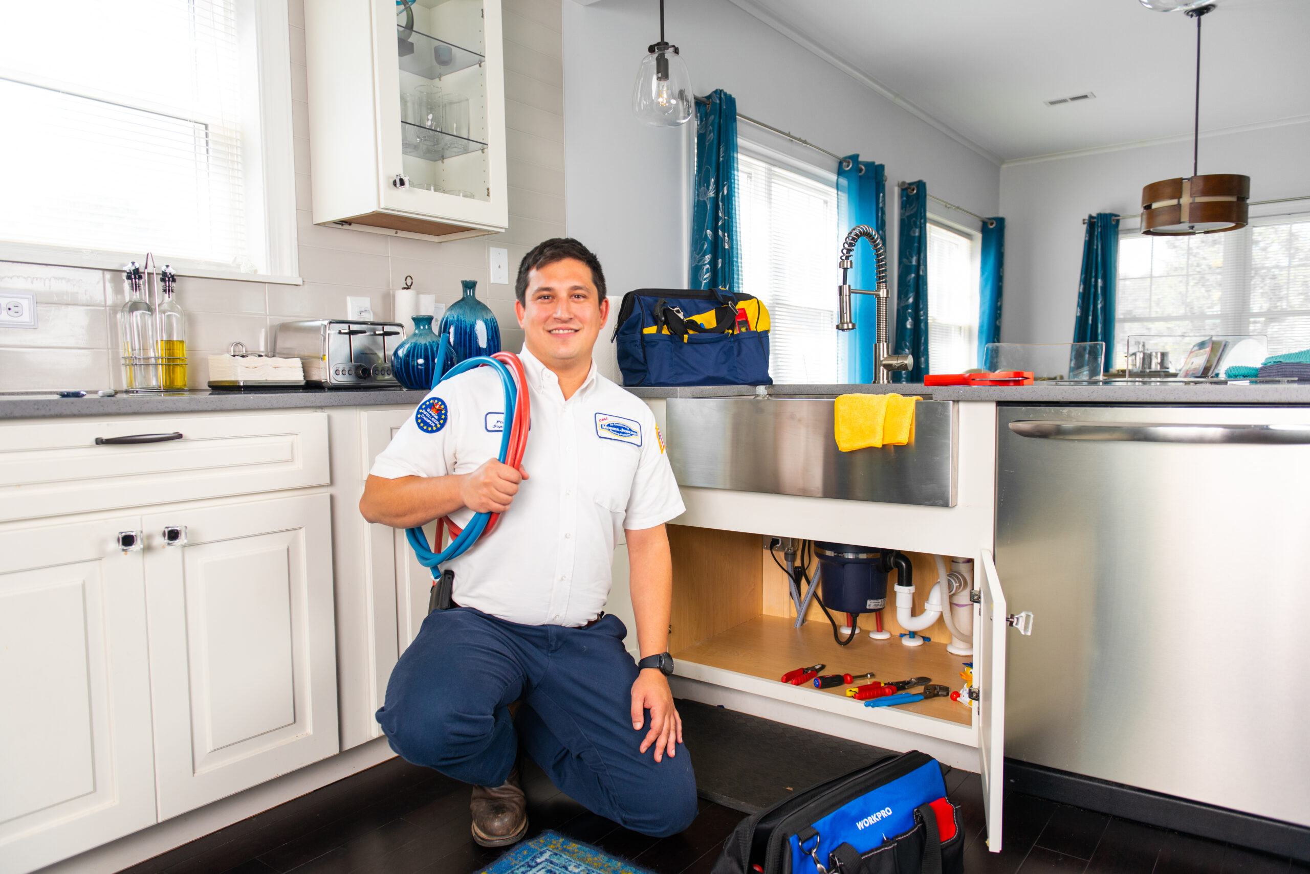 A service technician in a white uniform kneels in a kitchen with a coil of piping, posing next to an open under-sink cabinet showing plumbing.