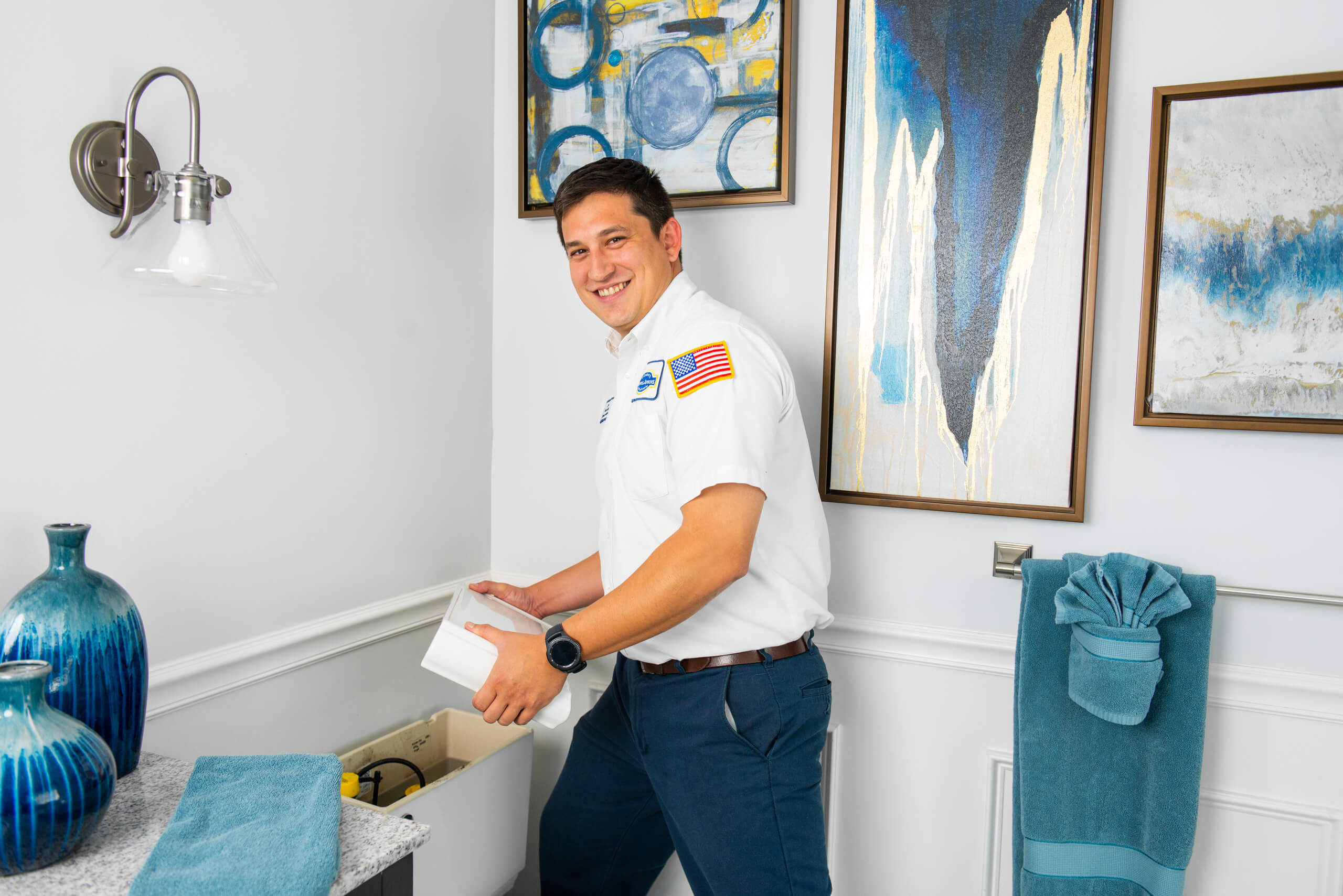 A plumber in uniform works on a toilet tank in a bathroom with blue decor and abstract artwork on the walls.