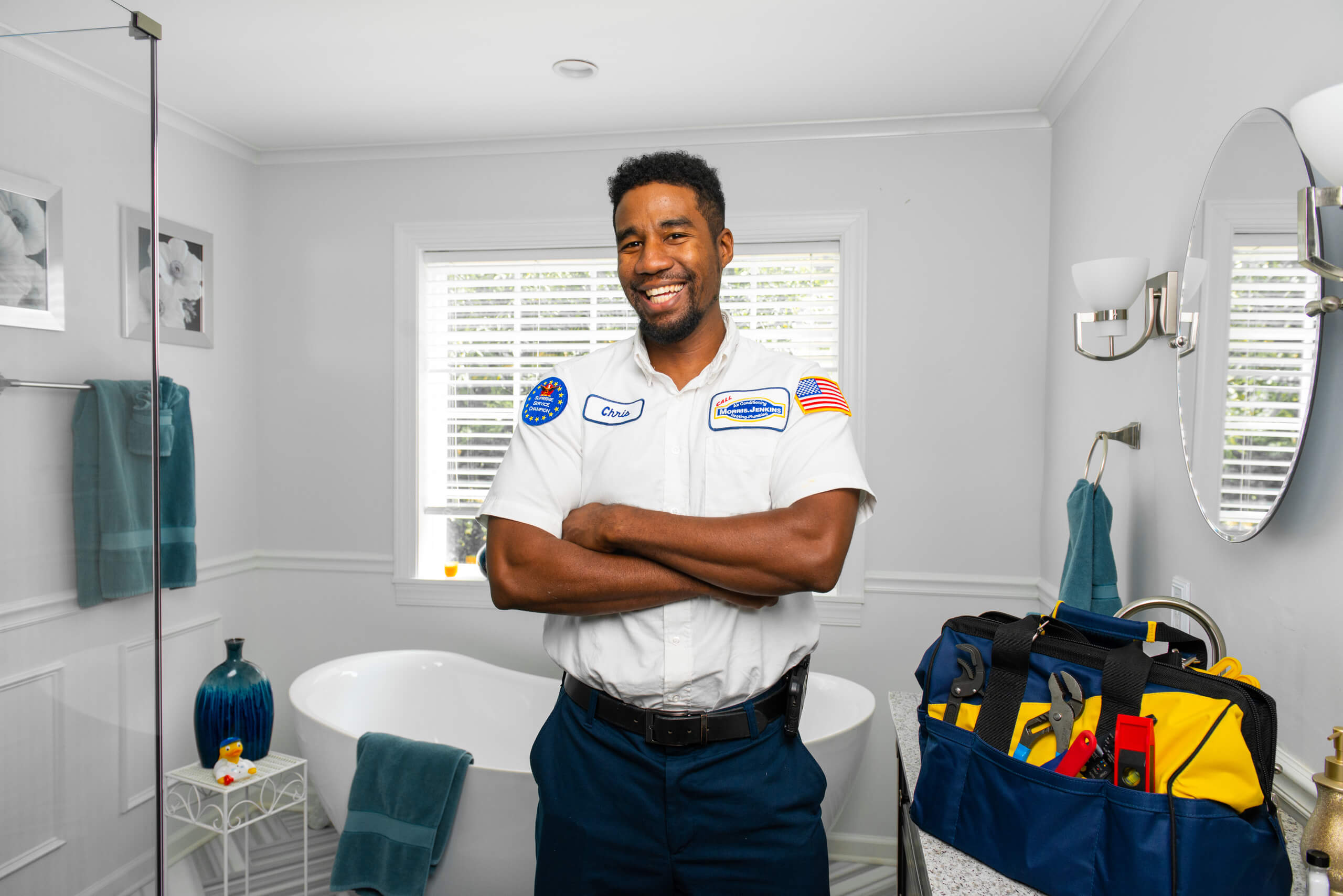 A smiling plumber in uniform stands with arms crossed in a bright bathroom, next to a tool bag on the counter.