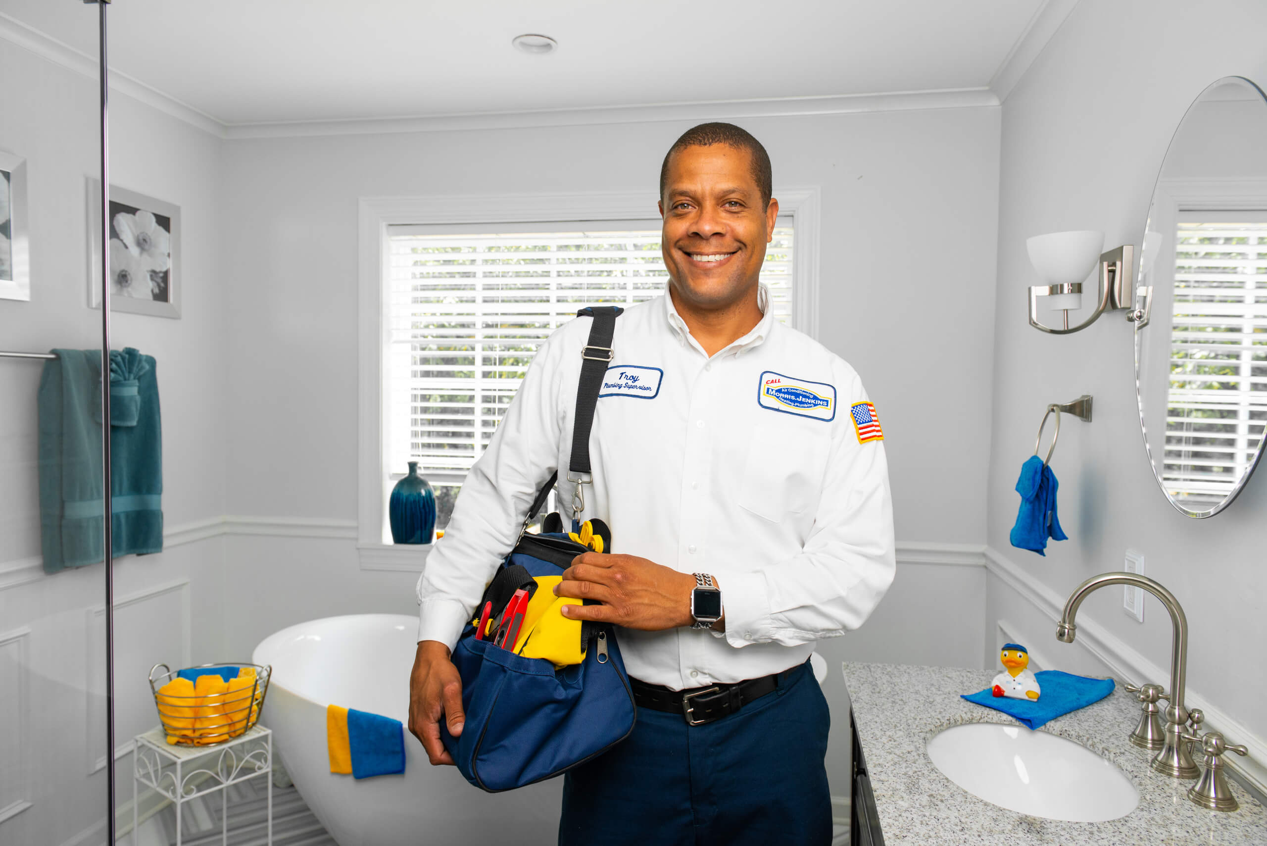 A plumber in uniform stands in a bright, clean bathroom, smiling and holding a tool bag near a sink and bathtub.