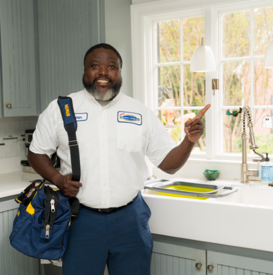 A plumber wearing a uniform stands in a kitchen by a sink, holding a tool bag and smiling while pointing toward the window.