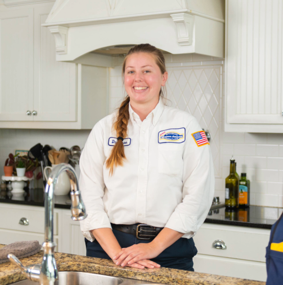 A plumber in uniform stands in a kitchen, smiling at the camera with hands folded on the counter.