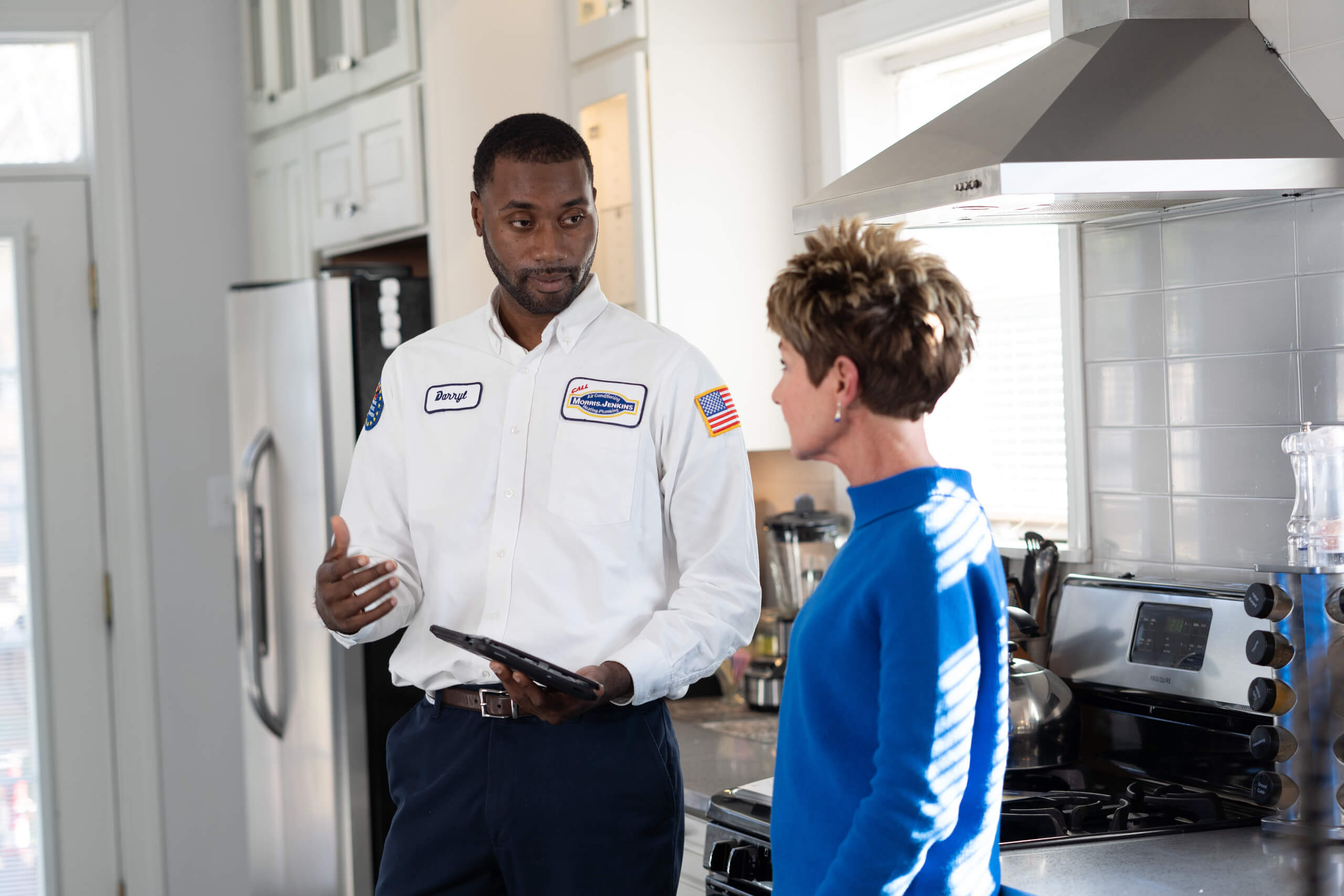 A technician in uniform holding a tablet talks to a woman in a kitchen next to a stove and range hood.